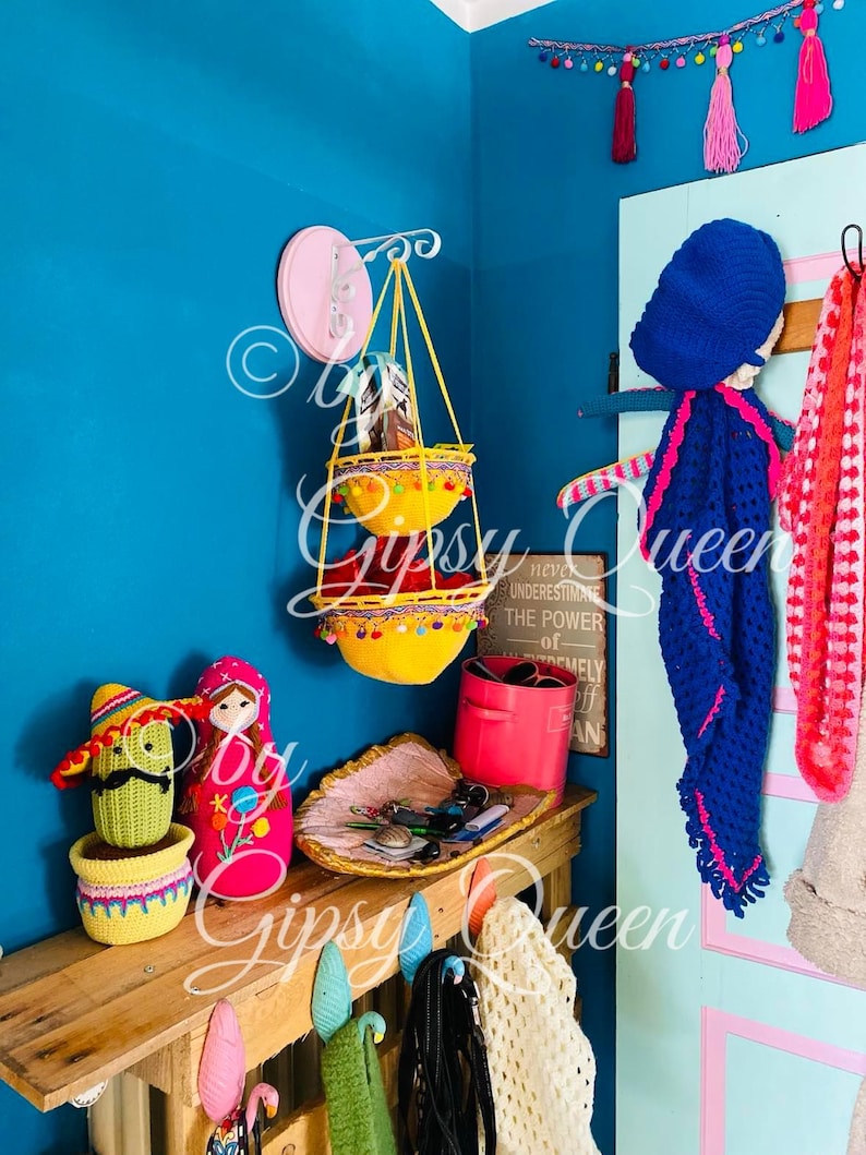 May include: A teal blue wall with a pink and white hanging wall organizer with two yellow crocheted baskets. The wall organizer is hanging from a white metal hook. There is a wooden shelf with a pink and white crocheted doll, a green crocheted cactus, a pink container, and a woven tray with keys and other small items. The shelf is decorated with a sign that reads "Never underestimate the power of extremely off plan".