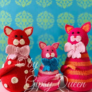 Op de afbeelding: Drie gehaakte katten speelgoed in rood, roze en oranje met witte stippen, roze en blauwe strikken en gestreepte patronen. Het speelgoed ligt op een roze oppervlak met een kleurrijke, gepatroneerde achtergrond. De tekst "By Gipsy Queen" is zichtbaar in de afbeelding.