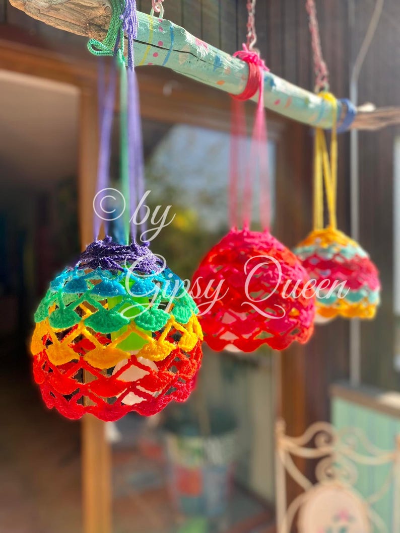 May include: Three crocheted rainbow-colored hanging ornaments with a white background. The ornaments are hanging from a driftwood branch painted turquoise with white polka dots. The text "by Gypsy Queen" is visible in the image.