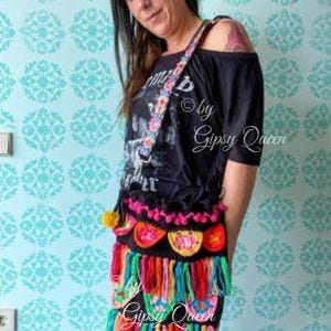 May include: A black crochet bag with colorful fringe and pom-poms. The bag has a floral patterned strap and is worn over the shoulder. The text "by Gipsy Queen" is visible on the bag.