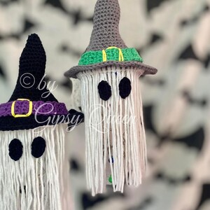 K&ouml;nnte beinhalten: Zwei handgefertigte Halloween-Geisterdekorationen. Jeder Geist hat einen wei&szlig;en K&ouml;rper mit schwarzen Augen und einen geh&auml;kelten Hexenhut. Ein Hut ist schwarz mit einem lila Band und einer goldenen Schnalle, der andere ist grau mit einem gr&uuml;nen Band.