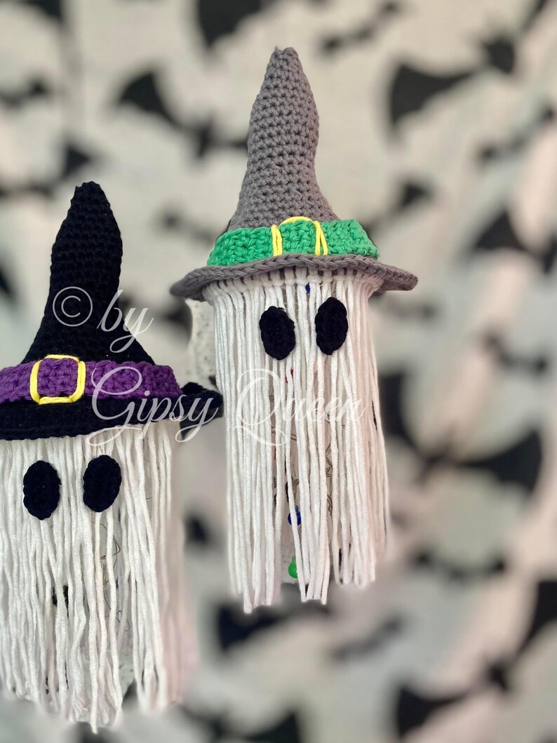 K&ouml;nnte beinhalten: Zwei handgefertigte Halloween-Geisterdekorationen. Jeder Geist hat einen wei&szlig;en K&ouml;rper mit schwarzen Augen und einen geh&auml;kelten Hexenhut. Ein Hut ist schwarz mit einem lila Band und einer goldenen Schnalle, der andere ist grau mit einem gr&uuml;nen Band.