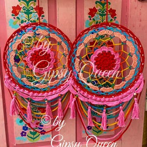 Könnte beinhalten: Zwei bunte gehäkelte Traumfänger mit rosa, roten, blauen, gelben und grünen Quasten, die an einer pinkfarbenen Wand mit Blumendesigns hängen. Die Traumfänger haben eine rote, weiße und blaue Mitte mit einem rosa und roten Außenring. Der Text "by Gipsy Queen" ist unten im Bild sichtbar.