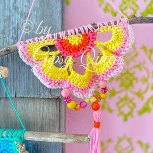 Pode incluir: Um pendurado de parede de croch&ecirc; em forma de flor com um esquema de cores rosa, amarelo e laranja. O pendurado est&aacute; preso a um galho com uma borla rosa e uma corda de contas.
