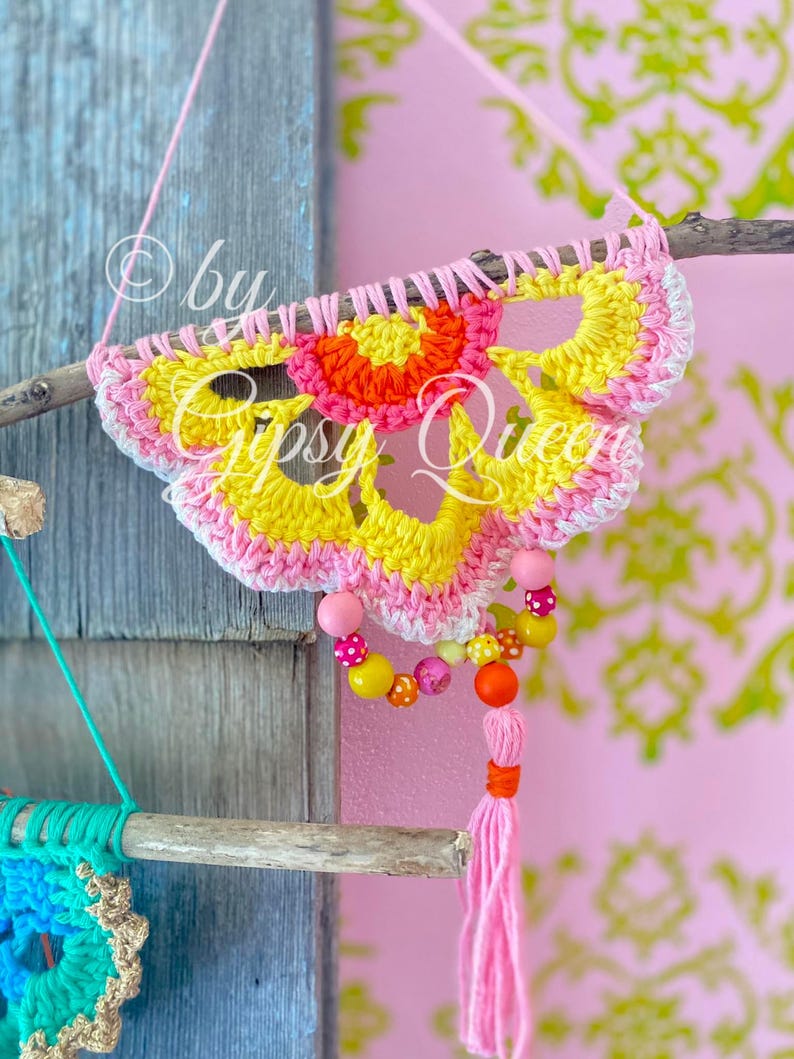Pode incluir: Um pendurado de parede de croch&ecirc; em forma de flor com um esquema de cores rosa, amarelo e laranja. O pendurado est&aacute; preso a um galho com uma borla rosa e uma corda de contas.