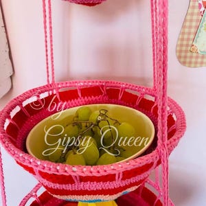 May include: A three-tiered hanging fruit basket made of crocheted yarn in pink, red, and blue. The top tier is empty, the middle tier holds green grapes in a white bowl, and the bottom tier holds bananas.