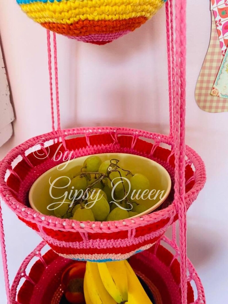 May include: A three-tiered hanging fruit basket made of crocheted yarn in pink, red, and blue. The top tier is empty, the middle tier holds green grapes in a white bowl, and the bottom tier holds bananas.