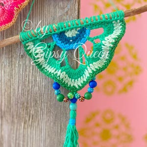 Pode incluir: Um pendurado de parede de croch&ecirc; verde, branco e azul com um tassel de contas. O pendurado &eacute; preso a um pau de madeira e tem um design circular no centro.