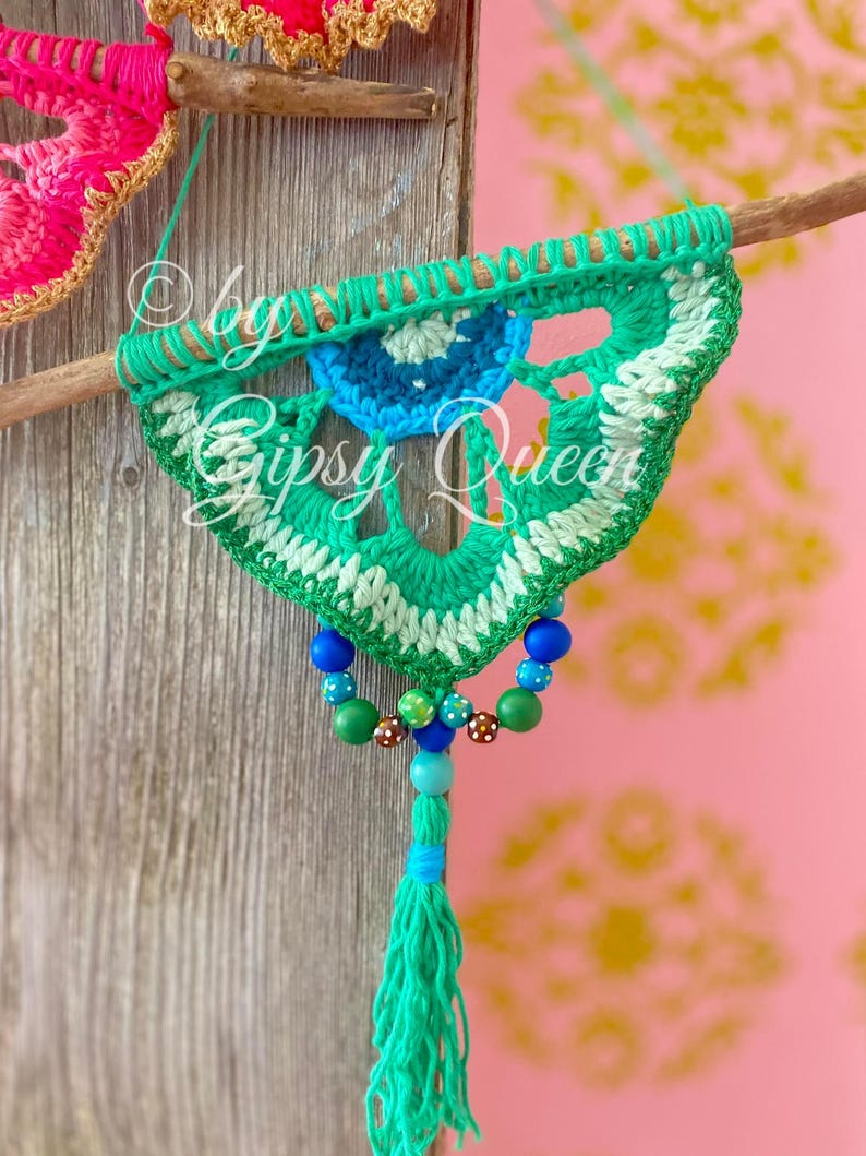 Pode incluir: Um pendurado de parede de croch&ecirc; verde, branco e azul com um tassel de contas. O pendurado &eacute; preso a um pau de madeira e tem um design circular no centro.