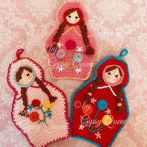 May include: Three crocheted hot pads shaped like Russian nesting dolls. Each doll is wearing a red hooded dress and has brown hair in braids. The dolls are decorated with colourful flowers and the text "by Gipsy Queen".