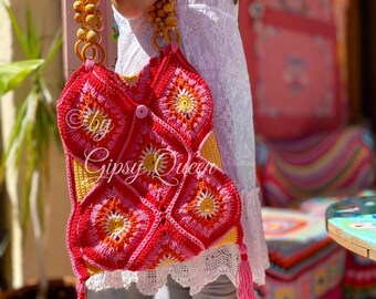 May include: A crocheted tote bag with a bright pink, red, and yellow floral pattern. The bag has a wooden handle and a tassel on the bottom. The bag is labeled "by Gipsy Queen".