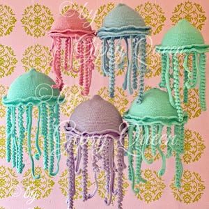 Puede incluir: Seis medusas hechas a mano con ganchillo en tonos pastel de rosa, azul y verde, colgadas sobre un fondo rosa con un diseño floral dorado. Cada medusa tiene una parte superior redondeada y tentáculos colgantes.