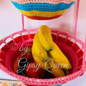 May include: Two crocheted hanging baskets, one pink and one with rainbow stripes, filled with bananas and other fruit. The pink basket has the text "&copy; by Gipsy Queen" on the side.