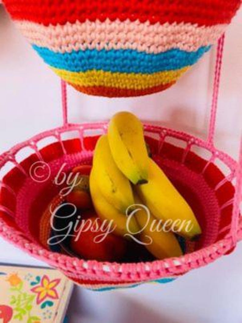 May include: Two crocheted hanging baskets, one pink and one with rainbow stripes, filled with bananas and other fruit. The pink basket has the text "&copy; by Gipsy Queen" on the side.