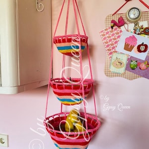 Könnte beinhalten: Drei Hängekörbe aus gehäkeltem Garn in Pink, Rot, Gelb und Blau. Die Körbe hängen an einem Metallhaken an einer rosa Wand. Der untere Korb enthält Bananen. Der Text "© by Gipsy Queen" ist in der unteren rechten Ecke sichtbar.
