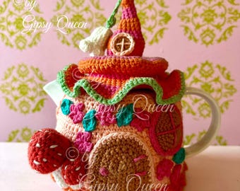 May include: A crocheted teapot cosy in the shape of a house with a red and orange roof, green trim, and a brown door. The cosy has a strawberry-shaped spout and the text "by Gipsy Queen" is visible on the side.