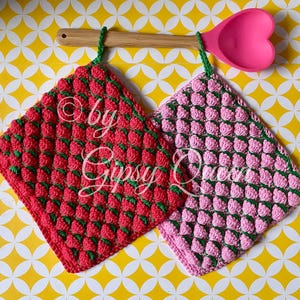 Puede incluir: Dos agarraderas de olla de ganchillo con forma de fresa. Una es roja con hojas verdes y la otra es rosa con hojas verdes. Están colgadas de una cuchara de madera con una cabeza de silicona rosa en forma de corazón.