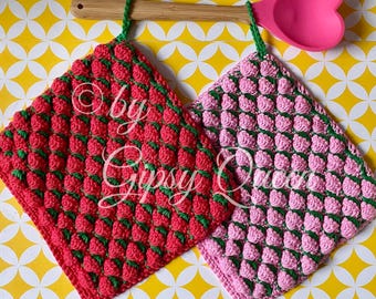 May include: Two crocheted pot holders shaped like strawberries. One is red with green leaves and the other is pink with green leaves. They are hanging from a wooden spoon with a pink heart-shaped silicone head.