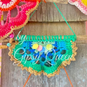 Pode incluir: Uma faixa de croch&ecirc; verde, azul e dourado com lantejoulas coloridas penduradas em um galho. A faixa tem o texto "Gipsy Queen".