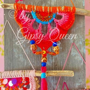 Pode incluir: Um tape&ccedil;aria de parede de croch&ecirc; colorida com um esquema de cores rosa, laranja e azul. A tape&ccedil;aria est&aacute; presa a um galho de madeira e apresenta uma franja de contas com contas coloridas. O texto "by Gipsy Queen" &eacute; vis&iacute;vel na imagem.