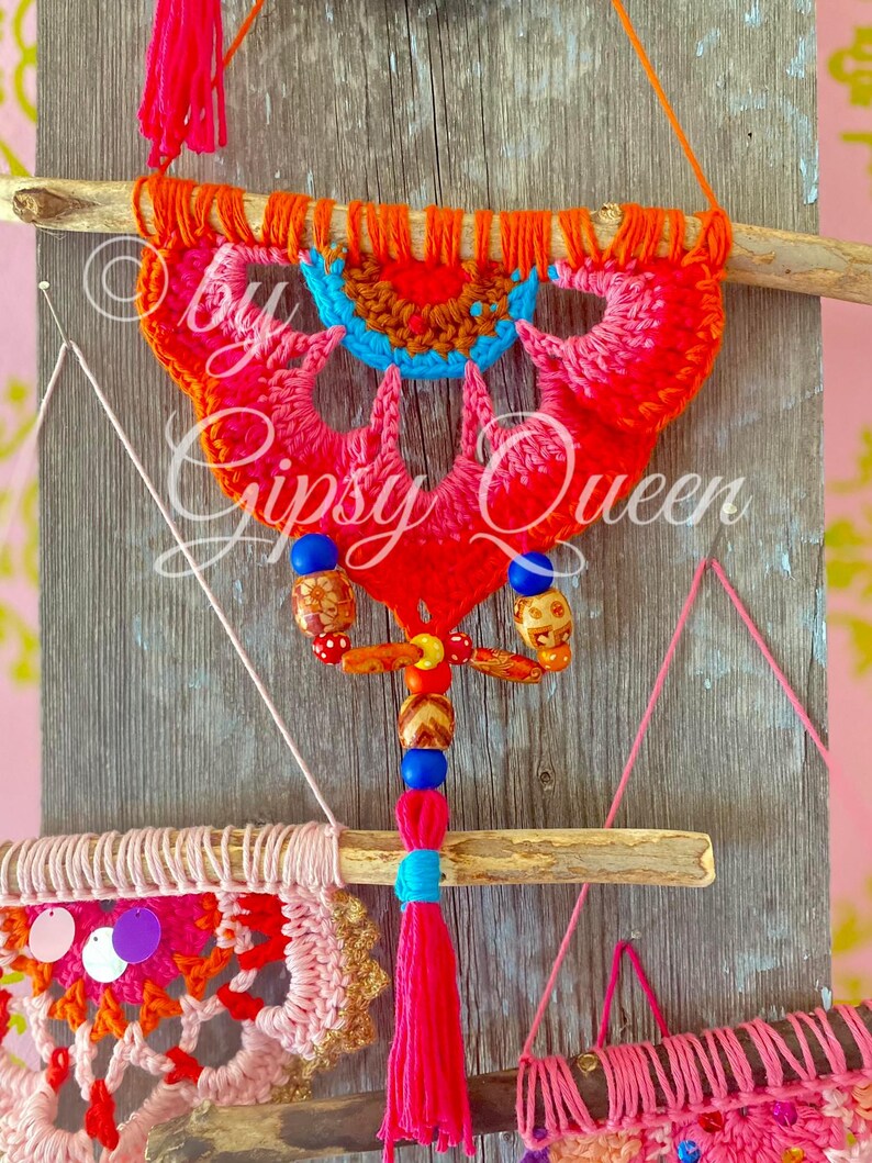 Pode incluir: Um tape&ccedil;aria de parede de croch&ecirc; colorida com um esquema de cores rosa, laranja e azul. A tape&ccedil;aria est&aacute; presa a um galho de madeira e apresenta uma franja de contas com contas coloridas. O texto "by Gipsy Queen" &eacute; vis&iacute;vel na imagem.