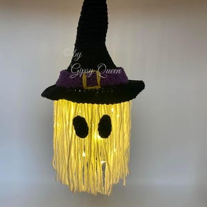 K&ouml;nnte beinhalten: Handgemachte Halloween-Dekoration in Form eines Geistes, mit einem schwarzen Hexenhut, einem lila Band und einer goldenen Schnalle. Das Gesicht des Geistes hat zwei schwarze ovale Augen, und der K&ouml;rper besteht aus gelbem Garn mit Innenbeleuchtung. Der Text "by Gipsy Queen" ist sichtbar.