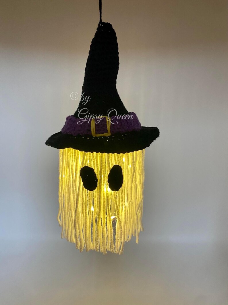 K&ouml;nnte beinhalten: Handgemachte Halloween-Dekoration in Form eines Geistes, mit einem schwarzen Hexenhut, einem lila Band und einer goldenen Schnalle. Das Gesicht des Geistes hat zwei schwarze ovale Augen, und der K&ouml;rper besteht aus gelbem Garn mit Innenbeleuchtung. Der Text "by Gipsy Queen" ist sichtbar.