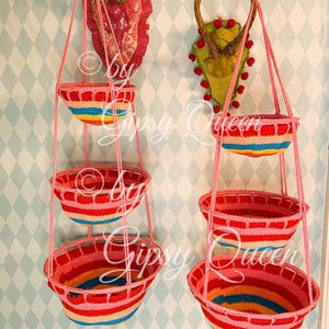 May include: Two sets of three hanging rainbow crocheted baskets. Each set is suspended from a pair of antlers mounted on a wall. The baskets are made of multi-colored yarn with pink, red, yellow, blue, and green stripes.