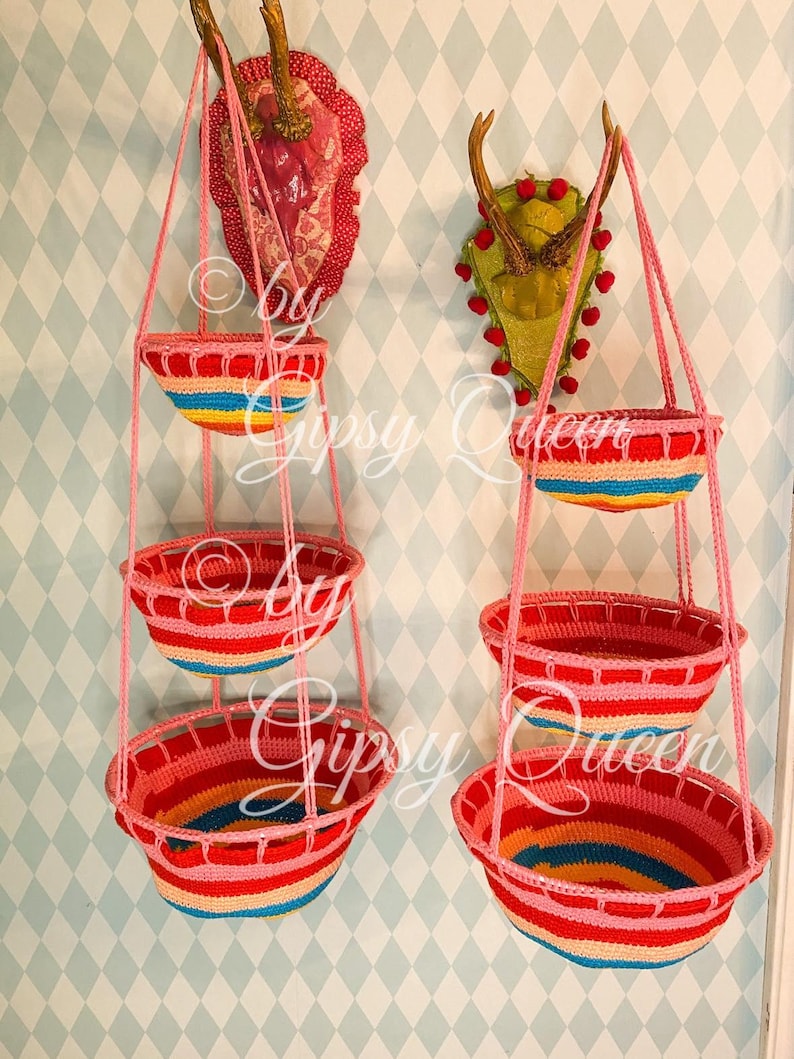 May include: Two sets of three hanging rainbow crocheted baskets. Each set is suspended from a pair of antlers mounted on a wall. The baskets are made of multi-colored yarn with pink, red, yellow, blue, and green stripes.