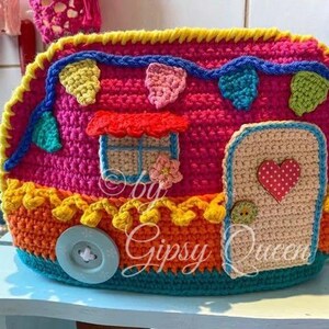 May include: A colorful, crocheted camper with a door, window, and button wheel. The camper is primarily pink, with accents of yellow, orange, blue, and green. The door has a red heart, and the window has a red awning.