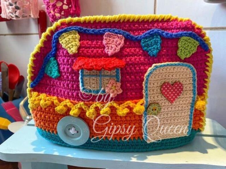 May include: A colorful, crocheted camper with a door, window, and button wheel. The camper is primarily pink, with accents of yellow, orange, blue, and green. The door has a red heart, and the window has a red awning.