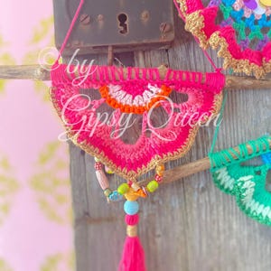 Pode incluir: Um pendurado de parede de croch&ecirc; rosa e laranja com um tassel de contas. O pendurado est&aacute; preso a um galho com uma borda dourada. O texto "by Gipsy Queen" &eacute; vis&iacute;vel no pendurado.