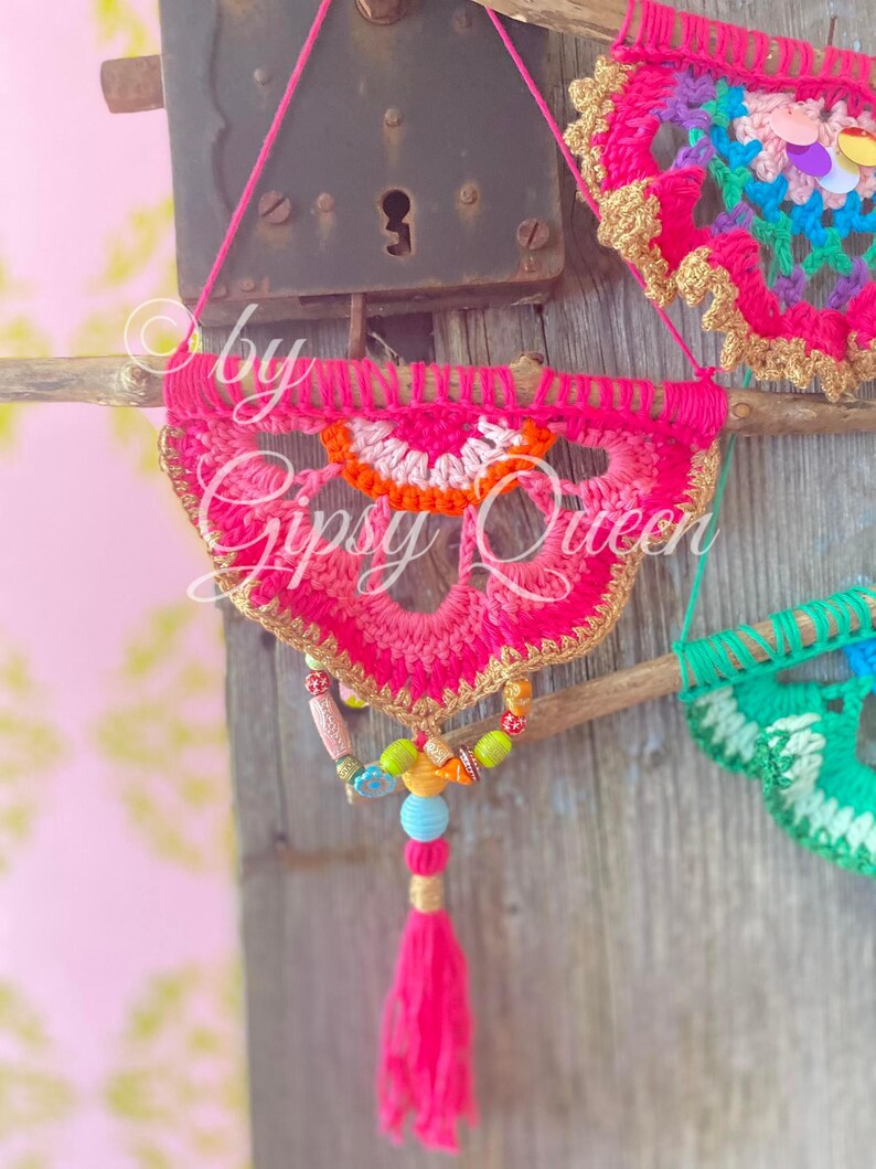 Pode incluir: Um pendurado de parede de croch&ecirc; rosa e laranja com um tassel de contas. O pendurado est&aacute; preso a um galho com uma borda dourada. O texto "by Gipsy Queen" &eacute; vis&iacute;vel no pendurado.