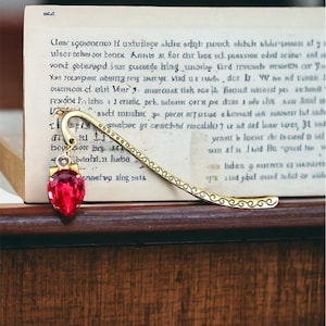 May include: A gold bookmark with a red glass heart charm. The bookmark is shaped like a long, thin ribbon with a curled end. The bookmark is lying on a book with old text.