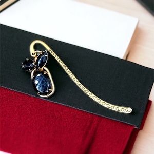 May include: A gold bookmark with a blue gemstone design. The bookmark is laying on top of a stack of books with black, white, and red covers.