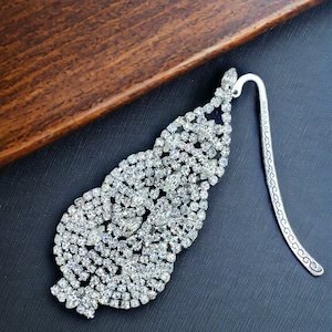 May include: A silver bookmark with a large, ornate design made of clear rhinestones. The bookmark is shaped like a leaf and has a long, thin tail.