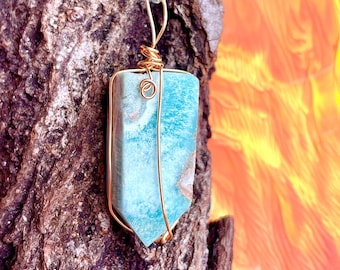 Pode incluir: Pingente com fio, apresentando uma pedra preciosa retangular azul clara com uma moldura de arame dourado. O pingente pende de um cordão trançado azul claro e branco. O fundo é um tronco de árvore texturizado e um design desfocado laranja e amarelo.