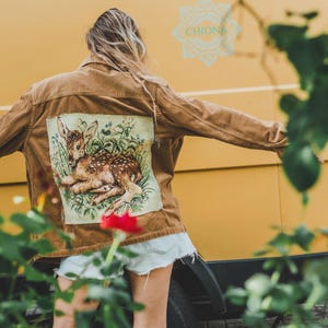 Könnte beinhalten: Eine beigefarbene Cordjacke mit einem großen rechteckigen Aufnäher auf dem Rücken, der ein detailliertes Bild eines im grünen Gras und zwischen Blumen ruhenden Rehkitzes zeigt. Die Jacke hat einen Kragen, lange Ärmel und eine Knopfleiste.