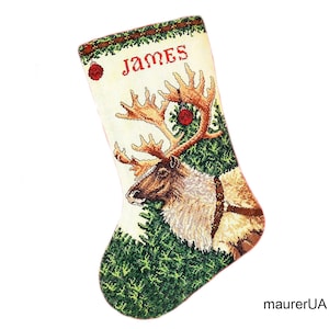 Puede incluir: Una media de Navidad blanca con un reno y ramas de pino verdes. La media está personalizada con el nombre "James" bordado.