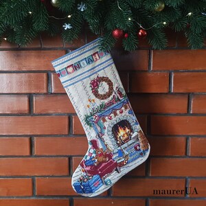 May include: A Christmas stocking with a festive fireplace scene, featuring a wreath, gifts, and a teddy bear. The name "NOAH" is embroidered at the top. The stocking is hanging on a brick wall, decorated with evergreen branches and ornaments.