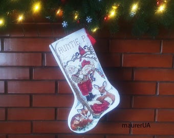 Könnte beinhalten: Weihnachtsstrumpf mit Kreuzstich-Design mit Weihnachtsmann, Hirschen und Füchsen in einer Winterszene. Der Strumpf trägt den Text "Auntie P" und eine rote Quaste. Er hängt an einer Backsteinmauer.