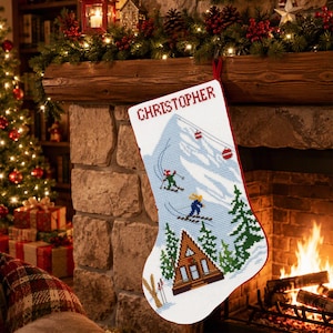 May include: A white Christmas stocking featuring a cross-stitch design of a snowy mountain, a brown A-frame cabin, green pine trees, and two skiers. The name CHRISTOPHER is stitched in red at the top. The stocking has a red trim and a red hanging loop.