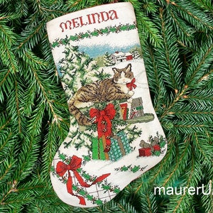 Puede incluir: Una media de Navidad blanca con un gato verde, rojo y marrón sentado encima de una pila de regalos. La media tiene el nombre "Melinda" bordado en la parte superior.