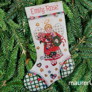 May include: White Christmas stocking with a red and green angel holding a teddy bear and a wreath. The stocking has the name "Emily Rose" embroidered on it.