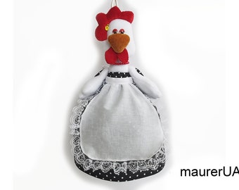 Puede incluir: Un porta-toallas de cocina con forma de pollo blanco con una cresta roja y un delantal de lunares negros y blancos. El porta-toallas tiene un borde de encaje blanco y un lazo para colgar.