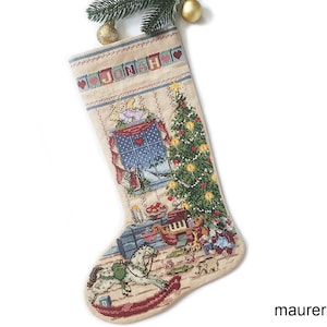 Può includere: Un calzino di Natale beige con un disegno ricamato di una finestra con un cielo blu, un albero di Natale e giocattoli. Il calzino ha il nome "Jenna" ricamato in alto.