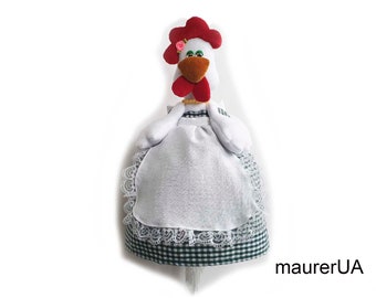 Puede incluir: Una figurita de pollo blanca con una cresta roja y un vestido a cuadros verdes y blancos. El pollo lleva un delantal blanco con ribete de encaje y tiene una flor rosa en la cabeza. El pollo sostiene un rollo de papel de cocina.