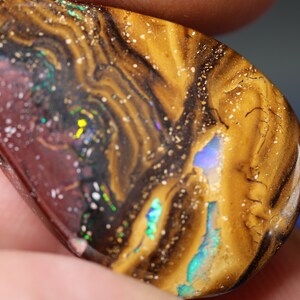 Könnte beinhalten: Ein brauner, schwarzer und goldener Opal-Edelstein mit schillernden Farbflecken. Der Opal hat eine Tropfenform und ist in einen silbernen Ring gefasst.