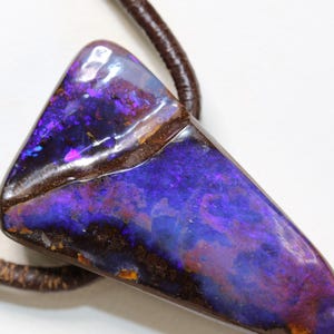 Könnte beinhalten: Ein polierter, dreieckiger Boulder-Opal-Anhänger mit tiefvioletten und blauen irisierenden Blitzen auf einer dunkelbraunen Eisensteinmatrix, der an einer dicken, strukturierten dunkelbraunen Lederbandkette hängt.