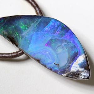 Könnte beinhalten: Ein schillernder Opal-Anhänger mit einer dunkelbraunen Lederschnur. Der Opal zeigt eine faszinierende Anordnung von Blau-, Grün- und Lilatönen. Der Edelstein ist in eine silberne Lünette gefasst. Ein einzigartiges Schmuckstück.